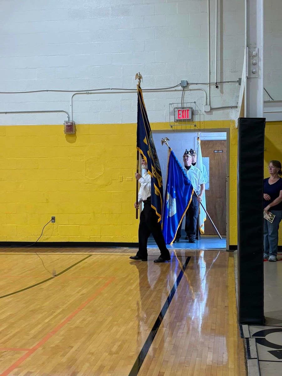 A Post 206 color guard carrying the United States flag and a service flag across a school gymnasium floor in front of a yellow cinder-block wall