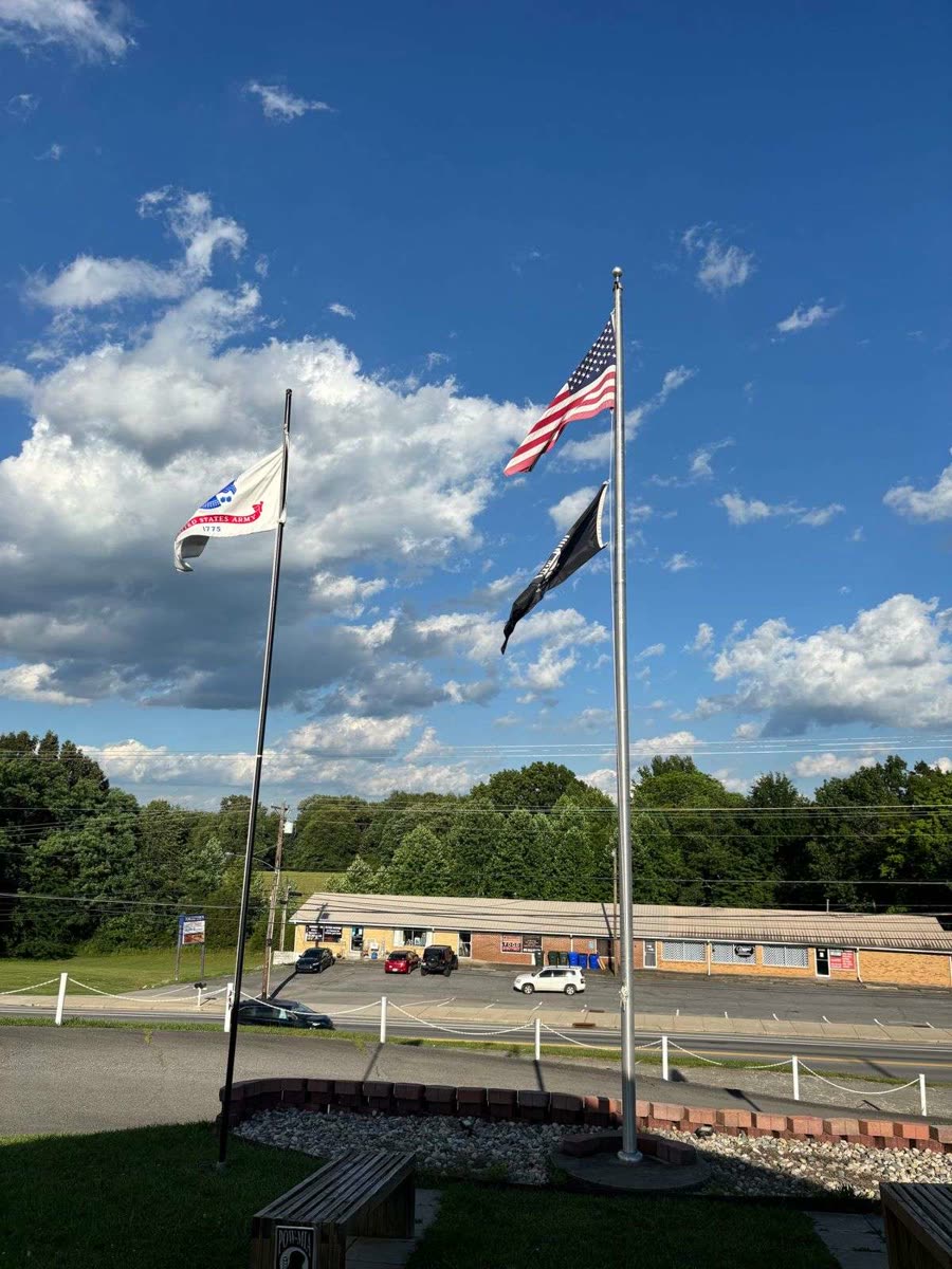 The United States flag flying above the black POW slash MIA flag on one pole, with a white United States Army flag on a second pole, against a blue sky with cumulus clouds
