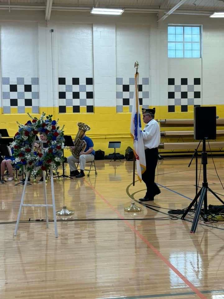 A memorial wreath on a stand beside a bugler playing at a ceremony in a school gymnasium