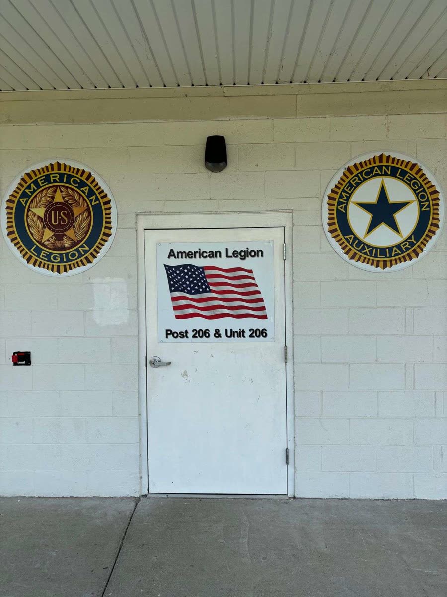 The entrance to the Post 206 building: a white door with a sign reading American Legion, Post 206 and Unit 206, flanked by an American Legion emblem plaque and an American Legion Auxiliary emblem plaque on a cream cinder-block wall