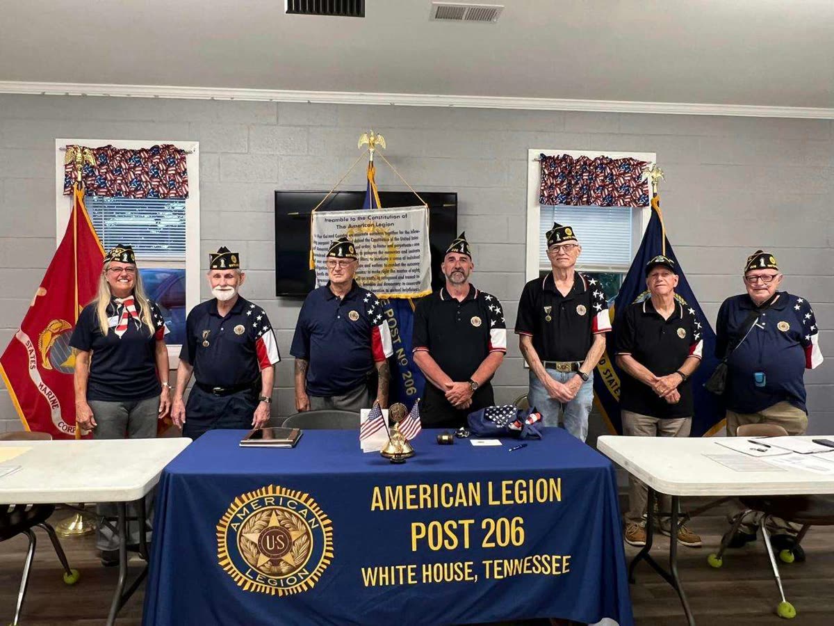 Seven members of Post 206 in American Legion caps standing behind a table draped with a banner reading American Legion Post 206, White House, Tennessee, with a Marine Corps flag and United States flags behind them
